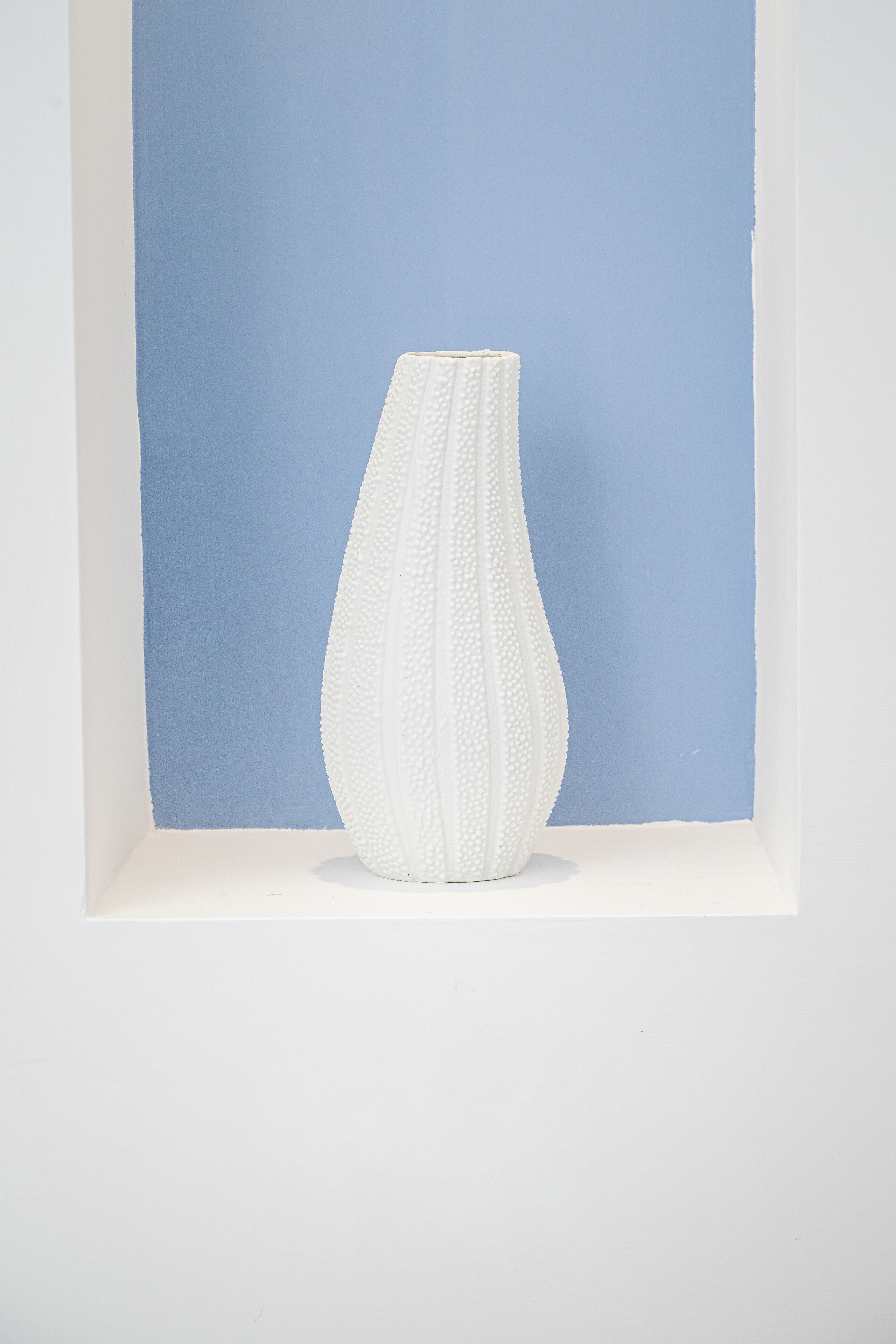 A white vase with a textured surface in a white-framed display against a blue background.