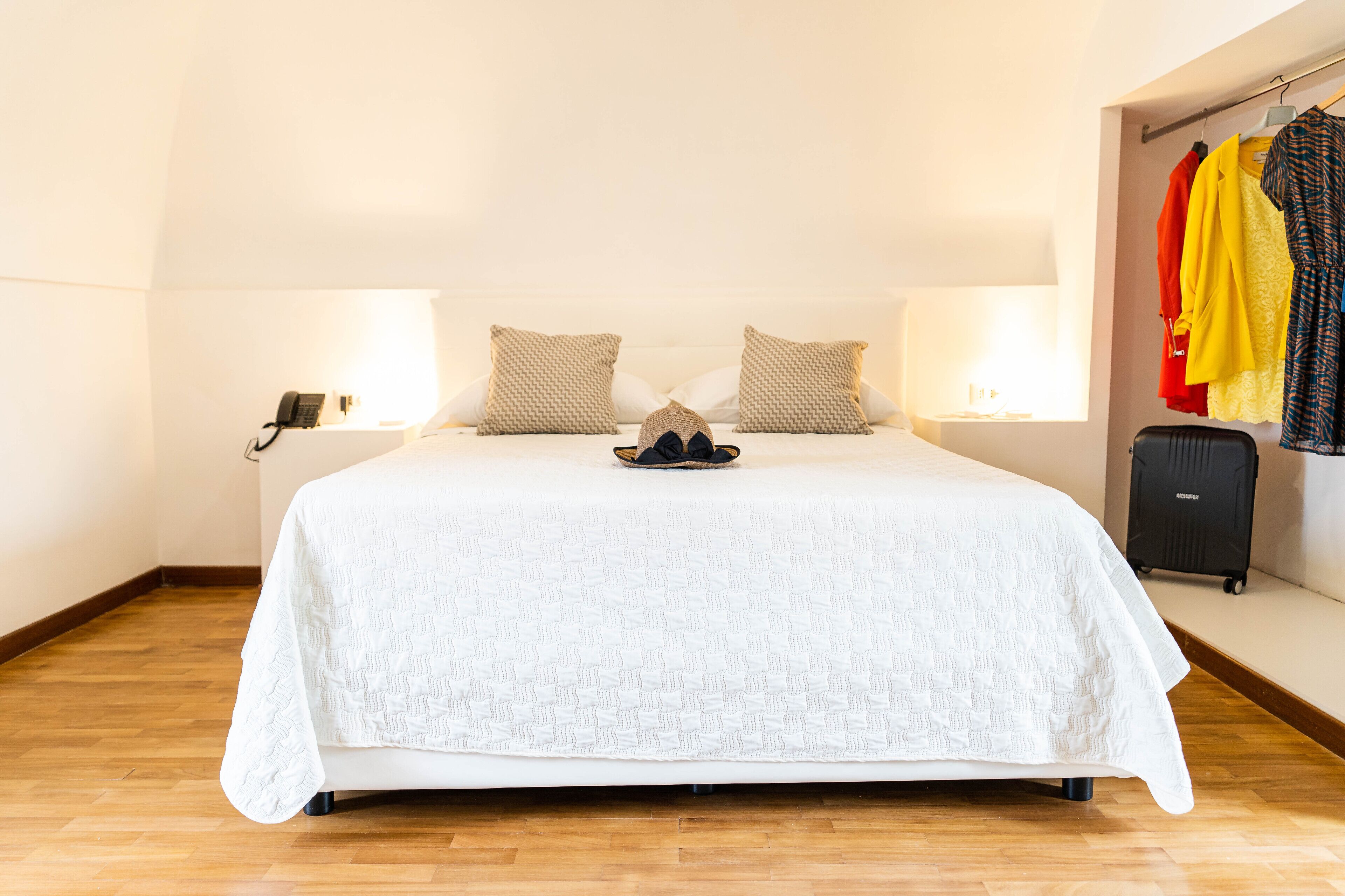 A neatly made bed with a white quilt, two beige pillows, and a pair of black sandals on the bed. There are two bedside lamps, a black suitcase on the floor, and a clothing rack with various garments.