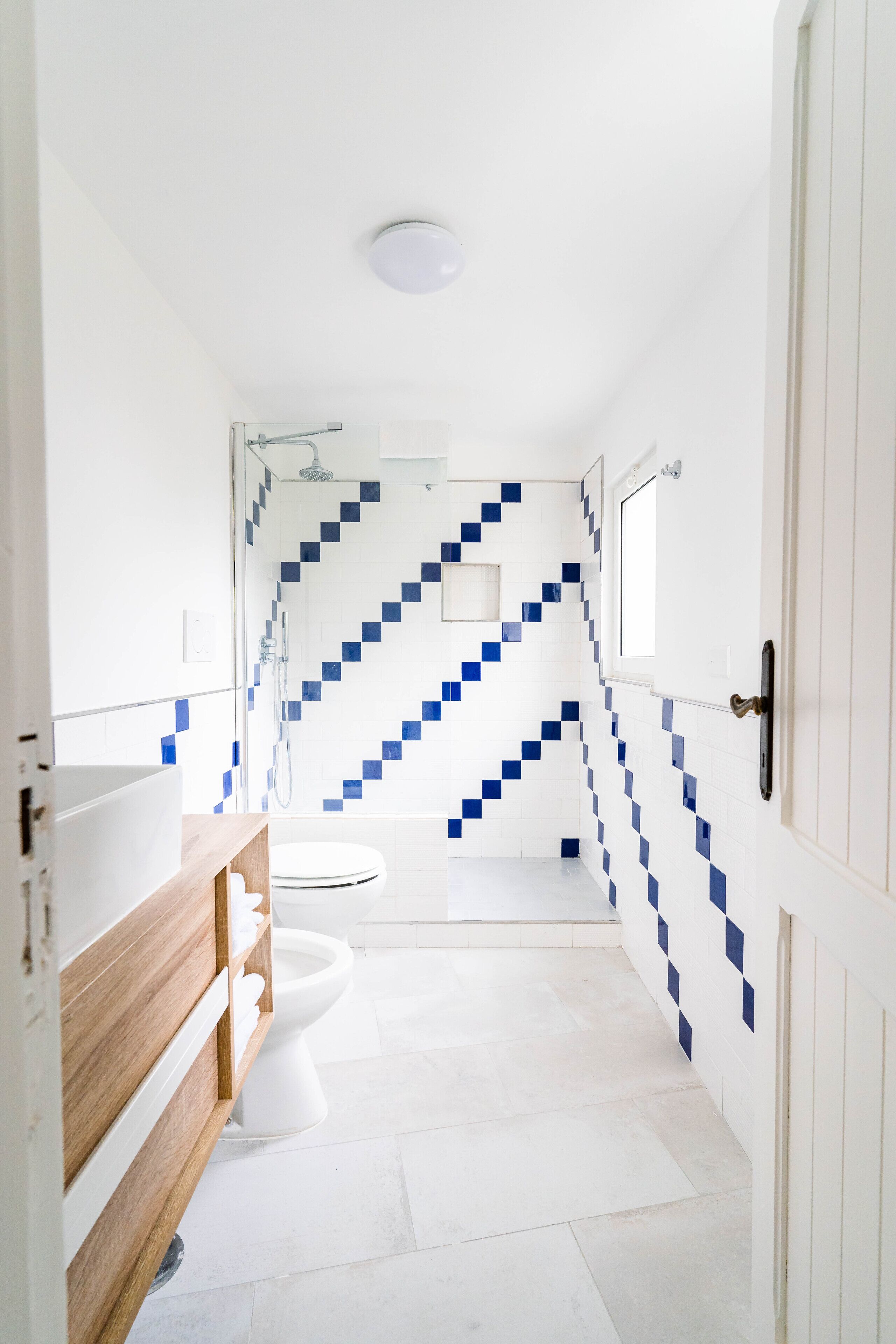 Un baño con lavamanos blanco, tocador de madera y zona de ducha con paredes de azulejos azules.