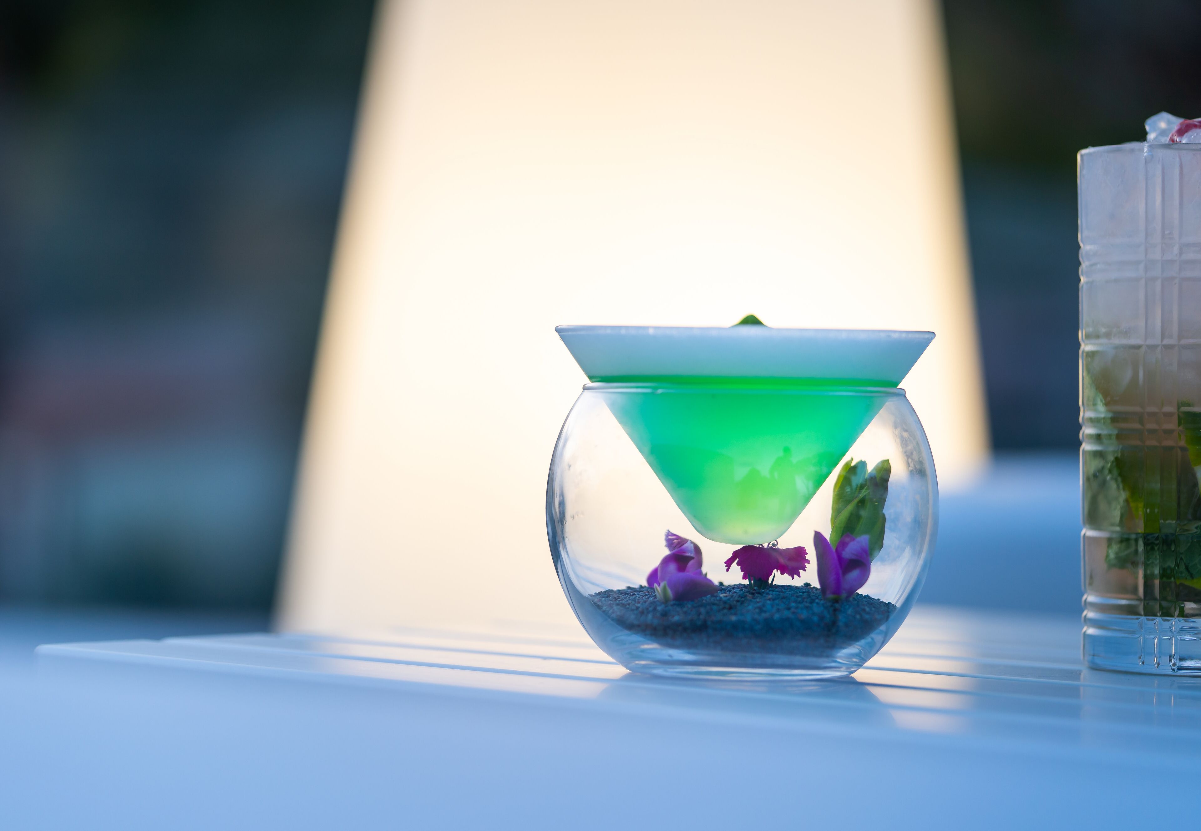 A clear glass bowl with a green liquid and purple flowers on a white surface.