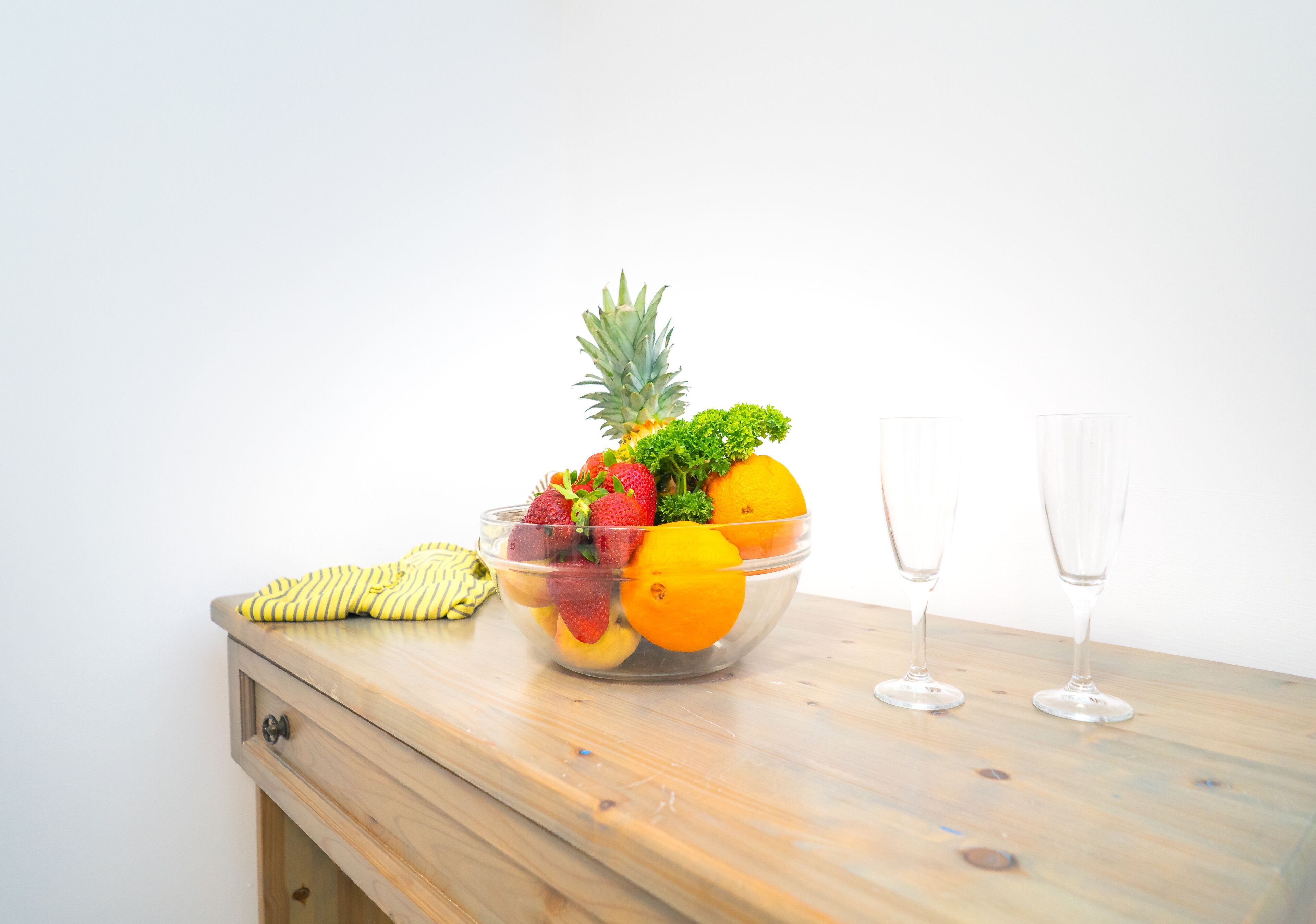 Een houten tafel met een fruitschaal met aardbeien, sinaasappels en een ananas, en twee lege wijnglazen.