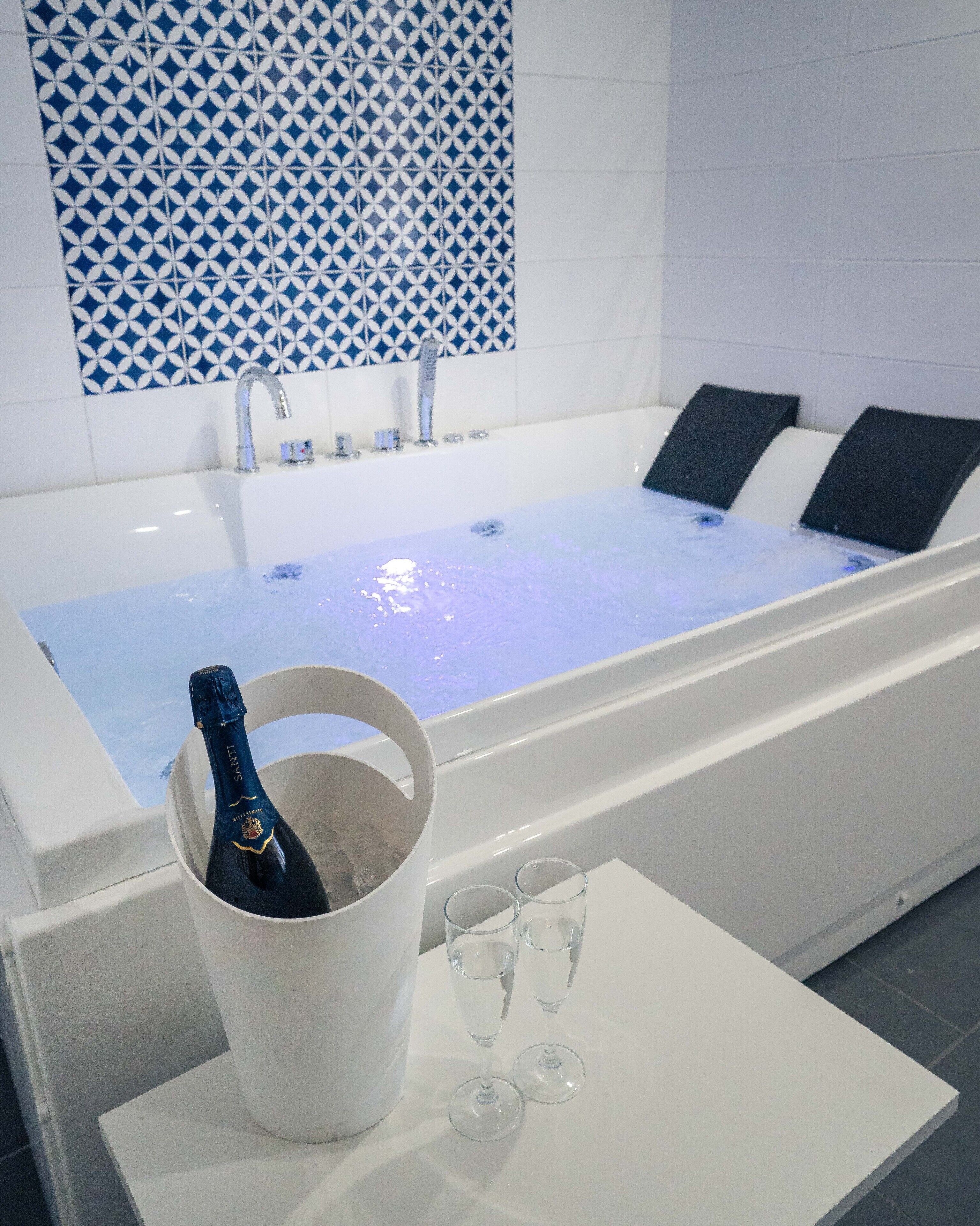 Una bañera con un papel tapiz azul estampado en la pared, una botella de champán en un cubo y dos copas sobre una mesa.