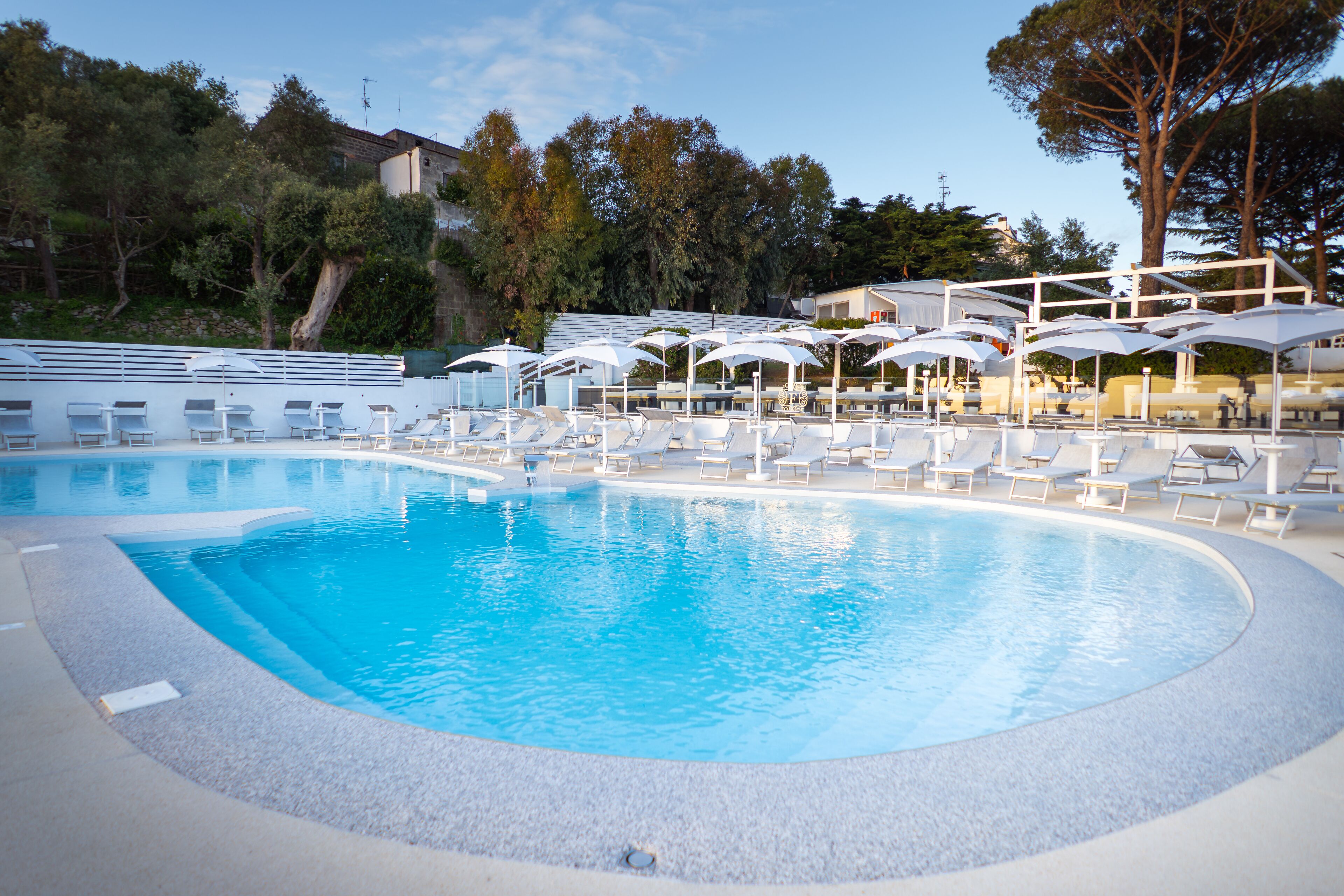 Piscina con sillas de descanso y sombrillas, rodeada de árboles y edificios.