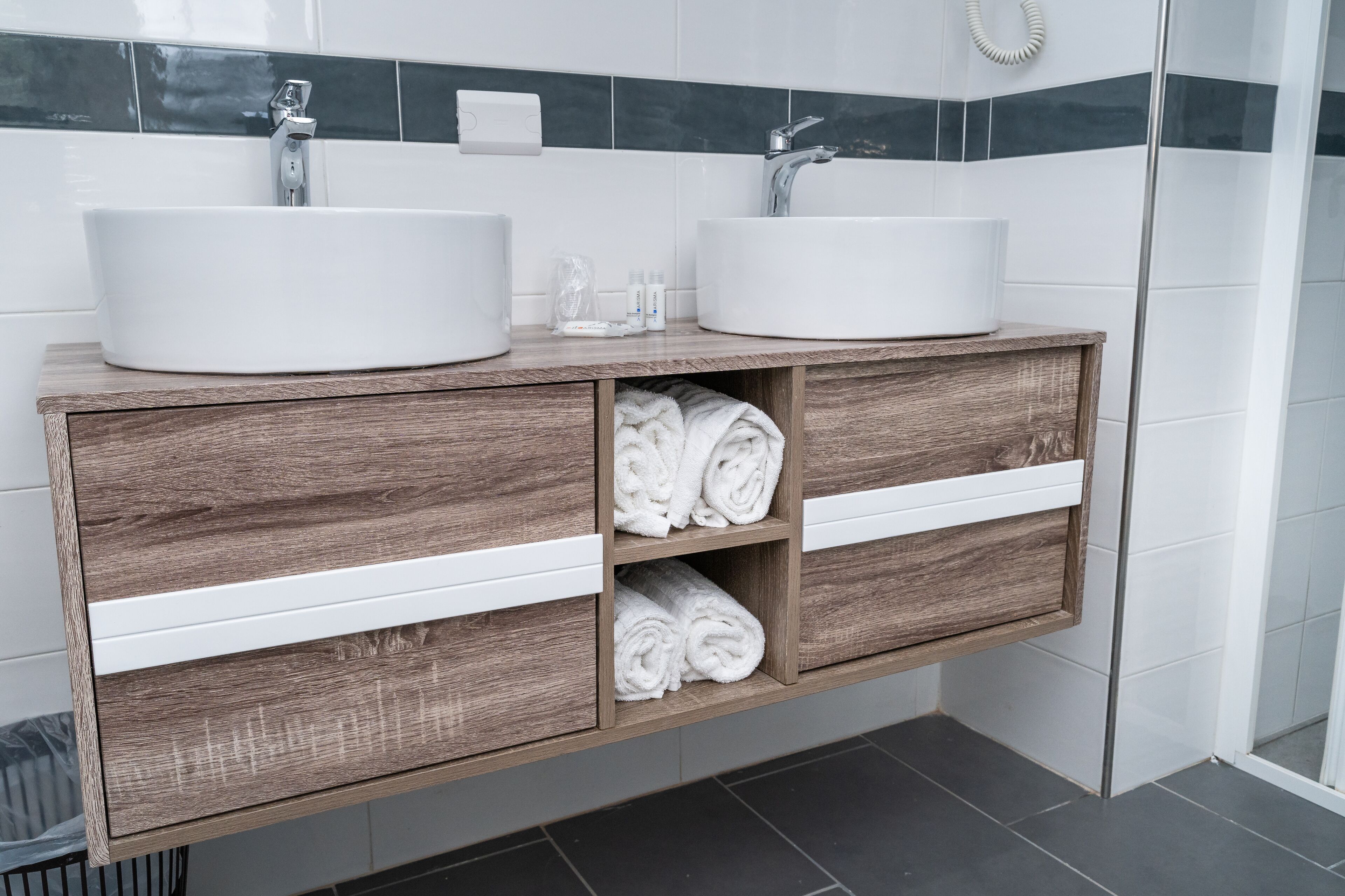 Un baño moderno con dos lavamanos blancos, tocador de madera y toallas enrolladas cuidadosamente.
