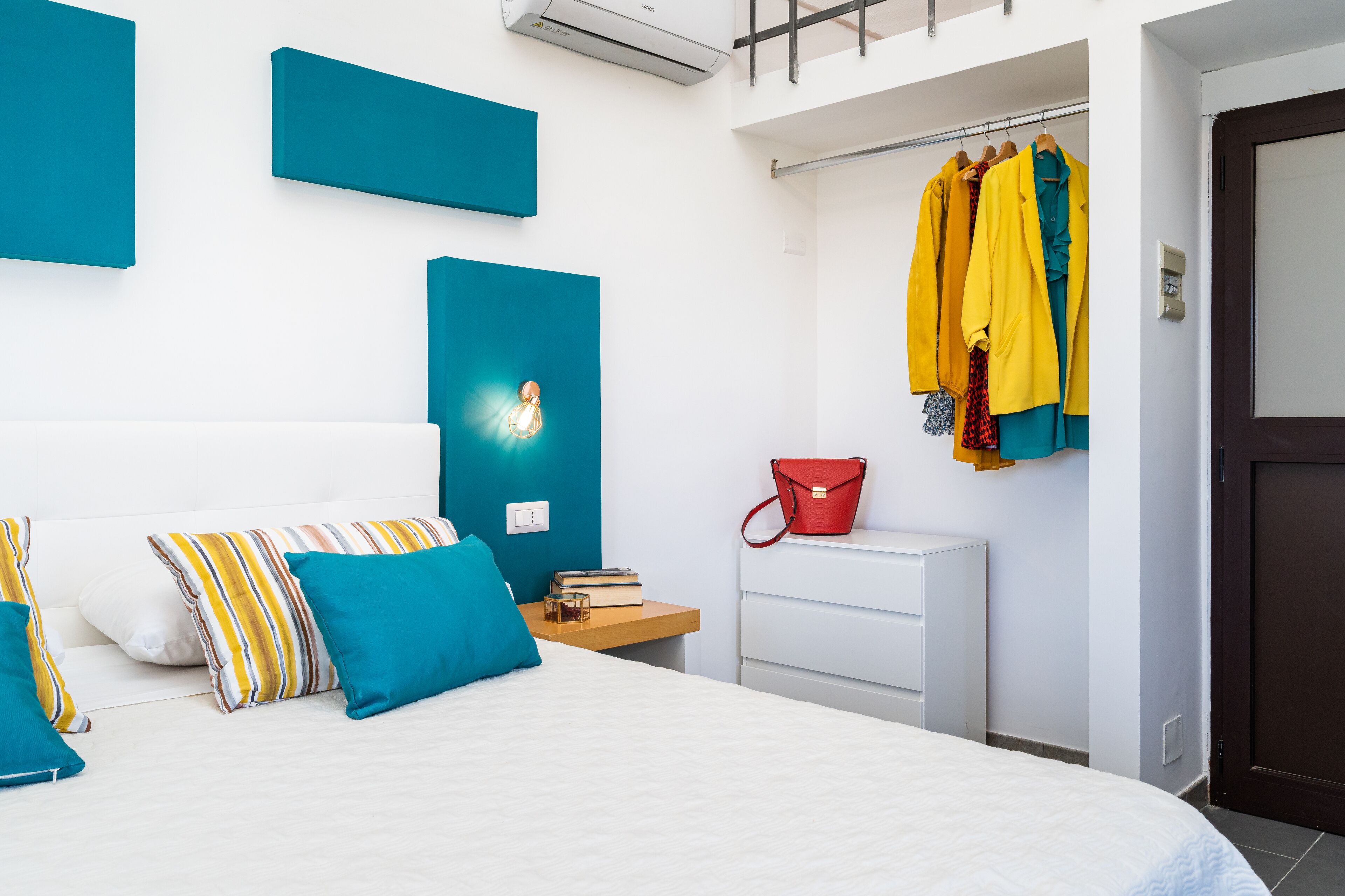 Une chambre moderne avec un lit, une table de chevet, un sac à main rouge et un porte-manteau avec des vestes jaunes et bleues.