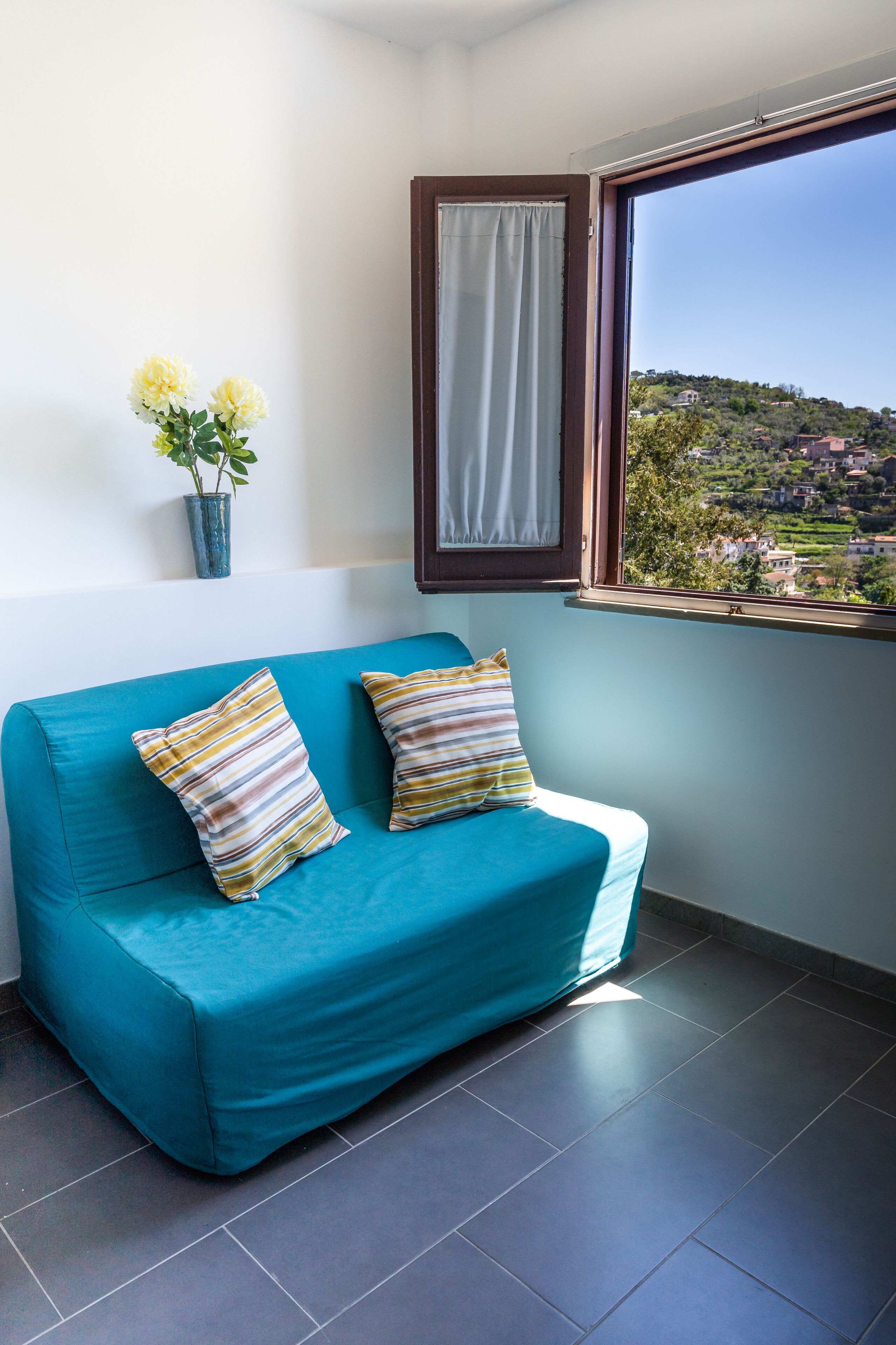 Un sofá color turquesa con almohadones a rayas, una planta en maceta y una ventana con vista a un paisaje montañoso.