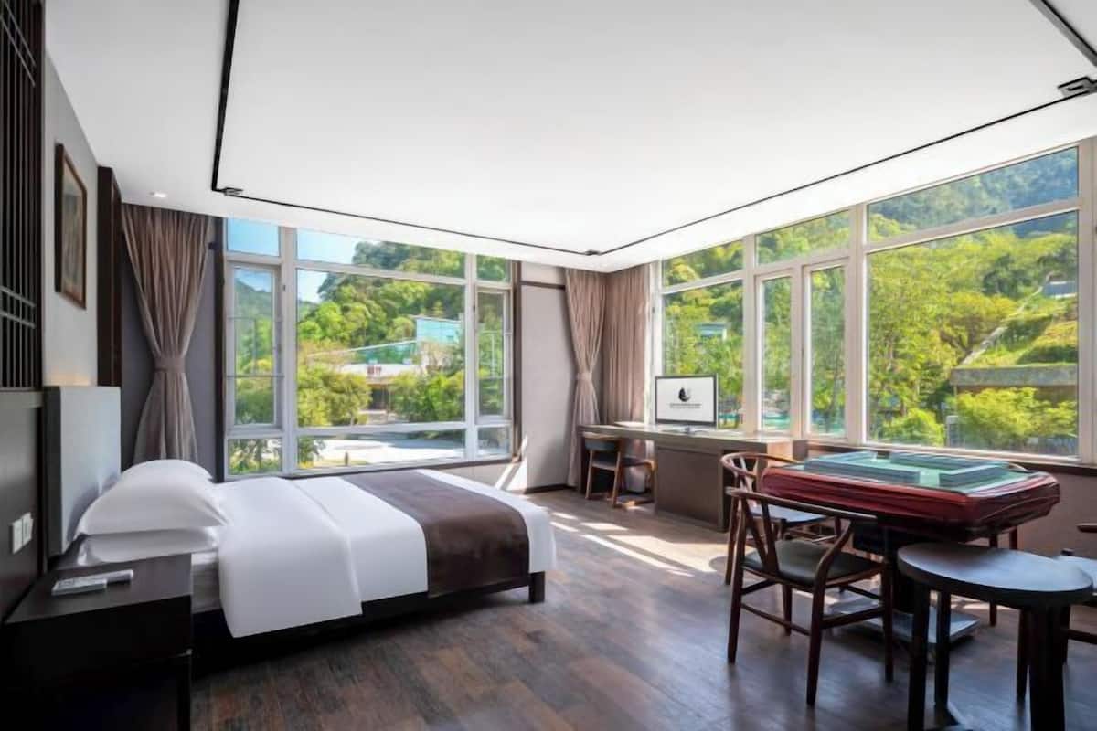 A modern hotel room with a large bed, a desk, and a view of greenery through large windows.