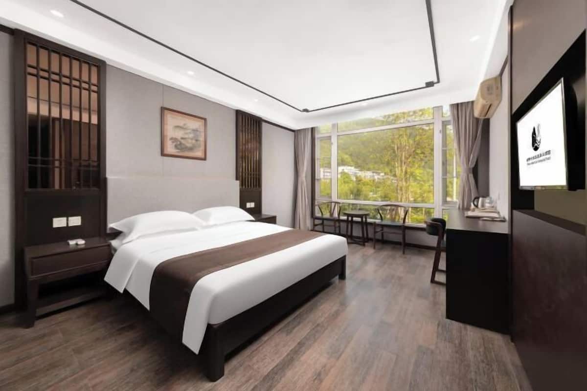 A modern hotel room with a large bed, a desk, and a view of greenery through the window.