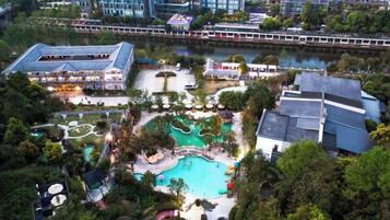 An aerial view of a resort with multiple swimming pools, a large building with a sloped roof, and a river in the background.