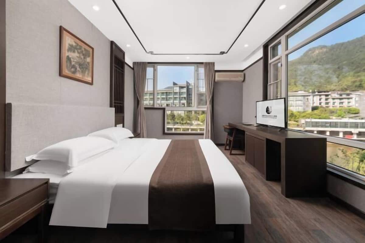 A hotel room with a large bed, a desk, a TV, and a balcony with a view of buildings and hills.