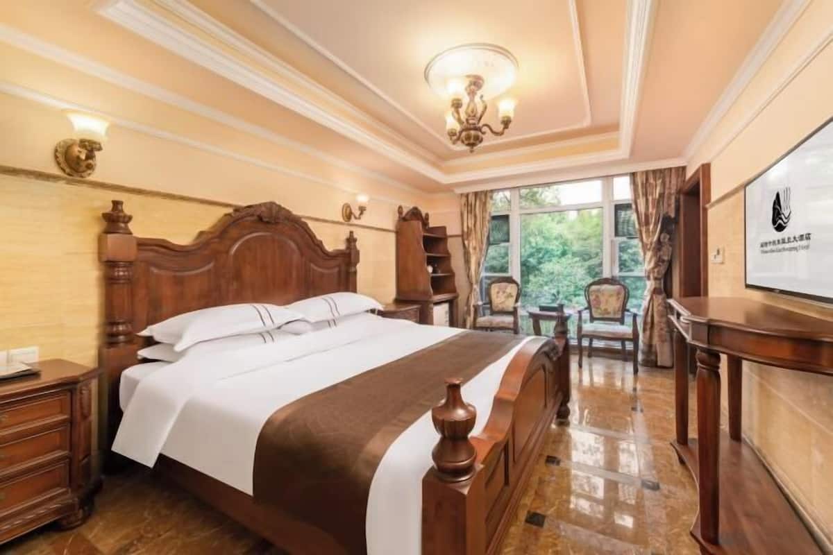 A hotel room with a large bed, a wooden headboard, a chandelier, a desk, and a view of greenery outside.