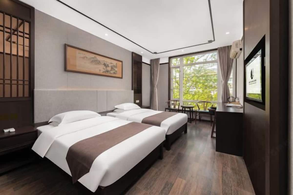 A hotel room with two beds, a desk, and a view of greenery outside.