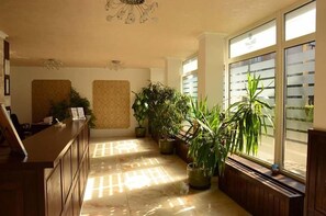 Vestíbulo de hotel con mostrador de recepción, plantas en macetas, una araña y ventanas amplias.