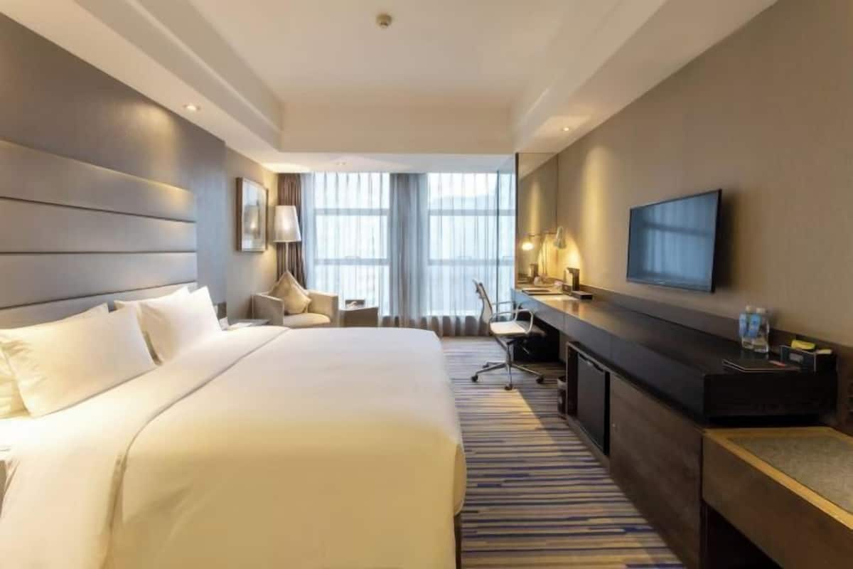 Une chambre d’hôtel avec un grand lit, un bureau avec une chaise, une télévision et une fenêtre avec des rideaux.