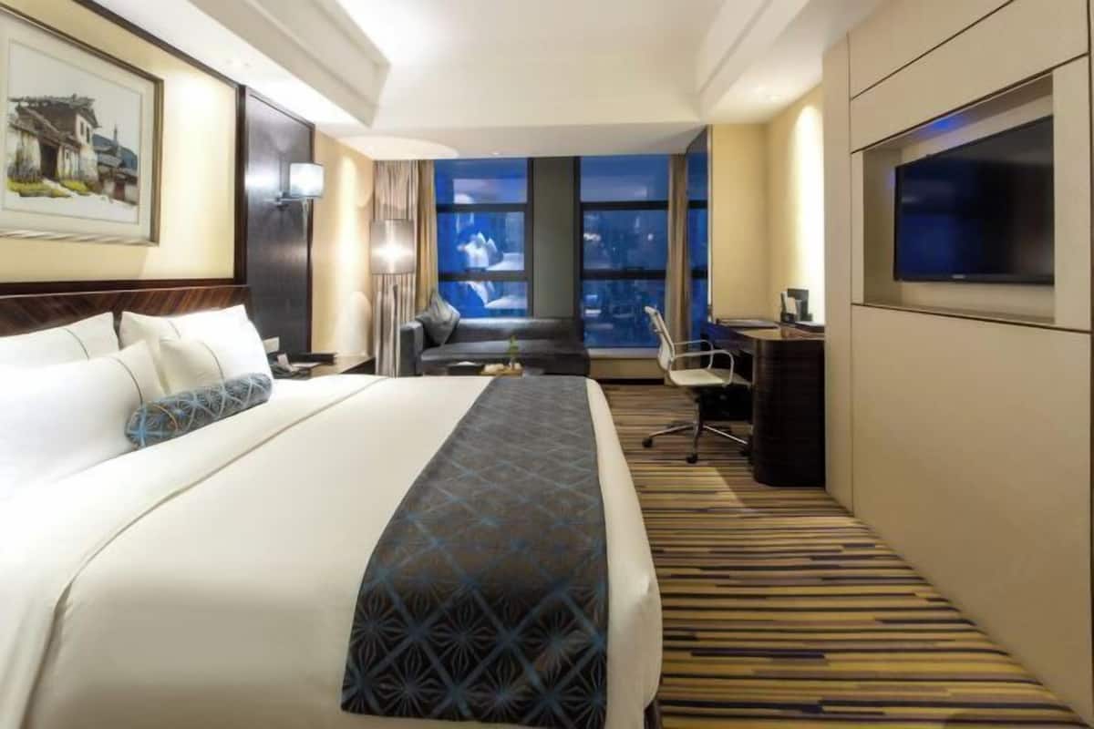 Une chambre d’hôtel avec un grand lit, un bureau, une chaise, une télévision et une vue sur la ville la nuit.