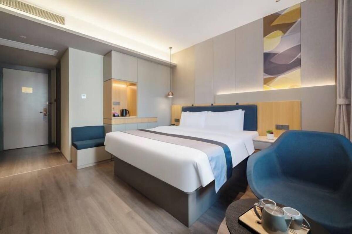 A modern hotel room with a large bed, a seating area, a desk, and a large window.