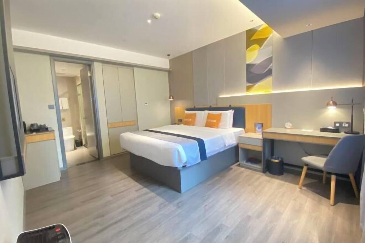 A modern hotel room with a large bed, a desk, and a chair. There is a wardrobe and a mirror on the wall.