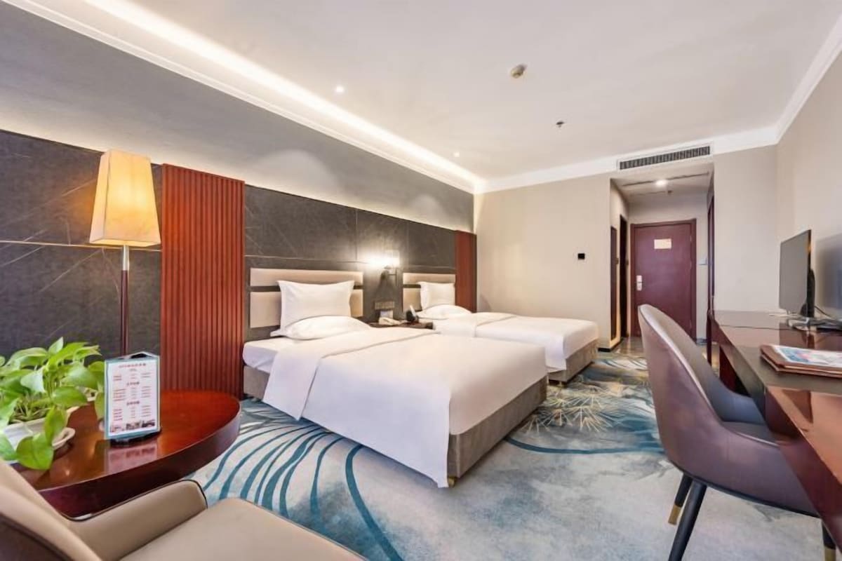 A hotel room with two beds, a desk, a chair, a lamp, and a plant.