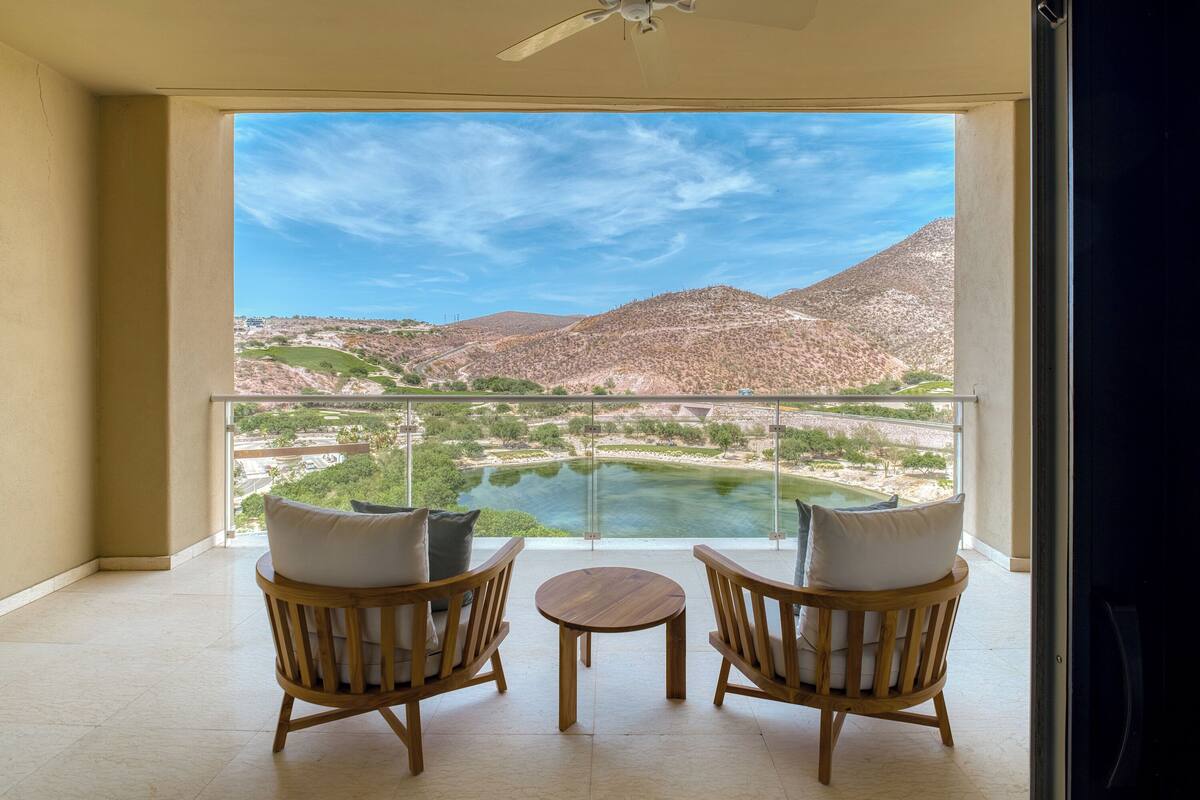 A balcony with two wooden chairs and a small table, overlooking a river and mountains.