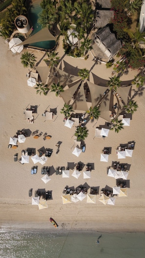 Une vue aérienne d’une plage avec de nombreux transats et parasols, des palmiers et une piscine.