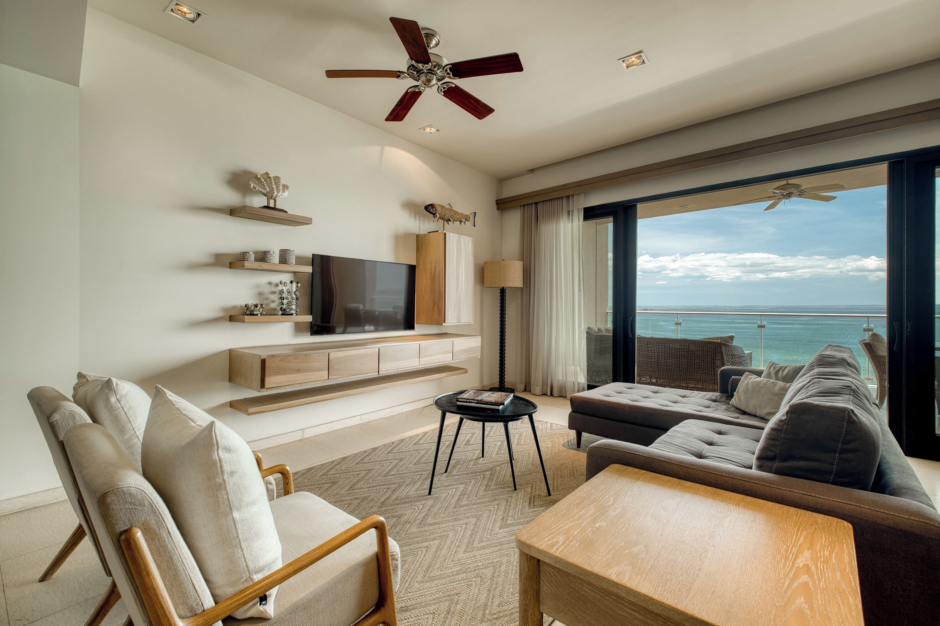 Ein modernes Wohnzimmer mit großem Fernseher, bequemen Sitzgelegenheiten, Deckenventilator und Meerblick.
