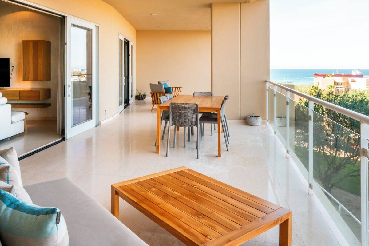A spacious balcony with a wooden table and chairs, a sofa, and a TV in the background.