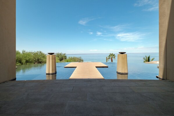 A infinity pool with a wooden deck leading to the water, surrounded by palm trees and a clear view of the ocean.