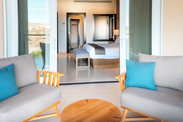 A modern hotel room with a large bed, two sofas, a wooden coffee table, and a view of the desert through large windows. - Puerta Cortés Residences by Lumina (La Paz)