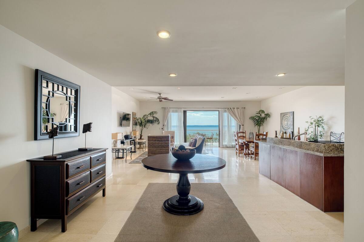 A spacious living area with a round table, a dark wooden cabinet, and a large window offering a view of the ocean.