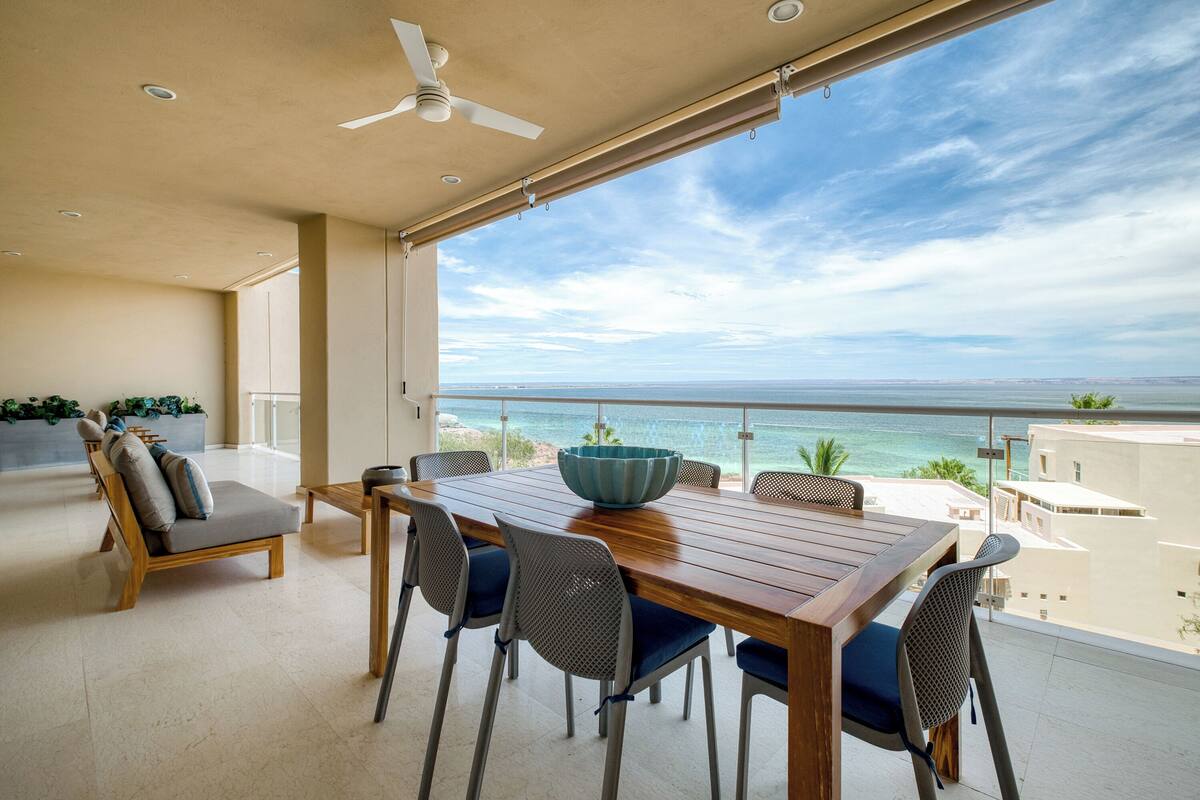 A dining area with a wooden table and chairs, a ceiling fan, and a view of the ocean.