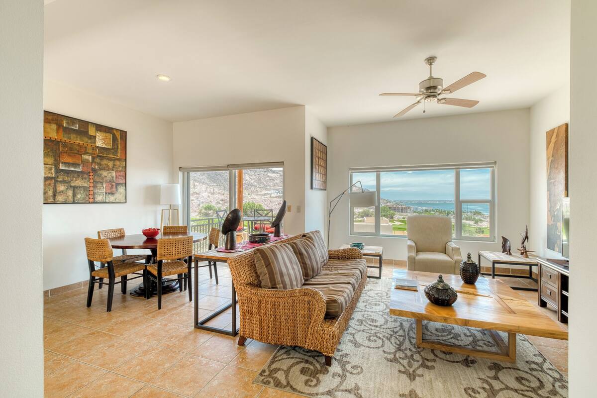 A spacious living room with a large sofa, dining table, and chairs. There is a ceiling fan, large windows with a view, and a rug on the floor.