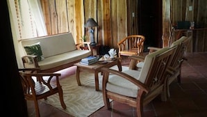 Een gezellige kamer met houten wanden, een bank, twee stoelen, een kleine tafel met een lamp en een raam met gordijnen.