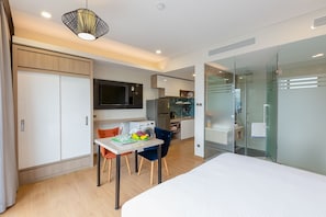 A modern hotel room with a bed, a dining area with a table and chairs, a kitchen area, and a glass-enclosed shower.