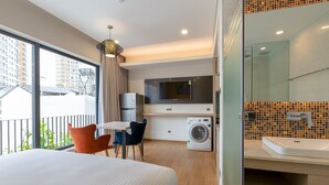 Une chambre d’hôtel moderne équipée d’un lit, d’un bureau avec des chaises, d’un lave-linge, d’une télévision et d’une salle de bain avec un lavabo et un miroir.