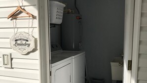 A laundry room with a washer and dryer, a small stool, a basket, and a sign indicating self-service laundry open 24 hours.