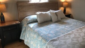A bedroom with a wooden headboard, a bed with a blue floral bedspread, two bedside tables with lamps, and a window with blinds.