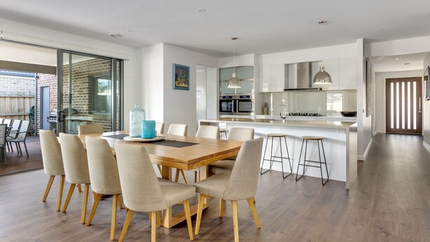 Et moderne køkken med en central ø, et spiseområde med et træbord og stole, samt en stor skydedør til en udendørs terrasse.