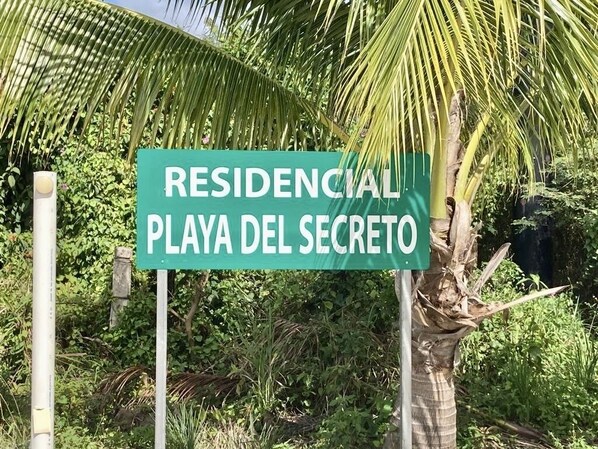 Une enseigne portant l’inscription « RESIDENCIAL PLAYA DEL SECRETO », placée dans un cadre naturel, avec de la végétation et un palmier.