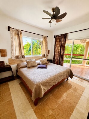 A bedroom with a bed, a ceiling fan, a window with curtains, a nightstand, and a view of greenery outside.