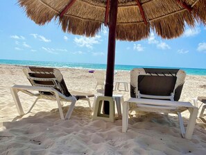 Two beach chairs under a thatched umbrella on a sandy beach with the ocean in the background.
