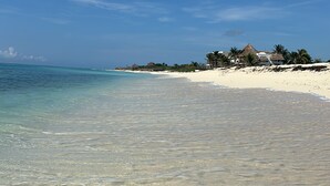 Una playa con aguas transparentes y poco profundas, una orilla arenosa y algunos edificios al fondo.