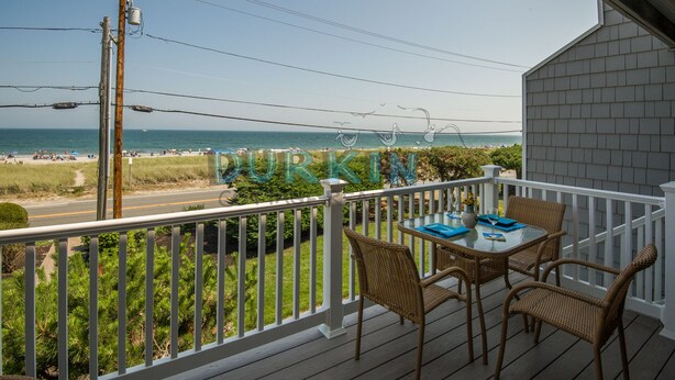 A balcony with wicker chairs and a table, overlooking a beach with a clear view of the ocean.