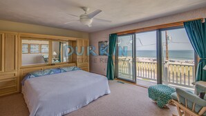 A bedroom with a large bed, a ceiling fan, a window with a view of the beach, and a wooden cabinet.
