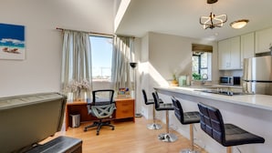 Een moderne keuken met witte kasten, een roestvrijstalen koelkast en een magnetron. Er is een houten bureau met een stoel en een kleine prullenbak. Een groot raam met gordijnen zorgt voor natuurlijk licht in de ruimte.