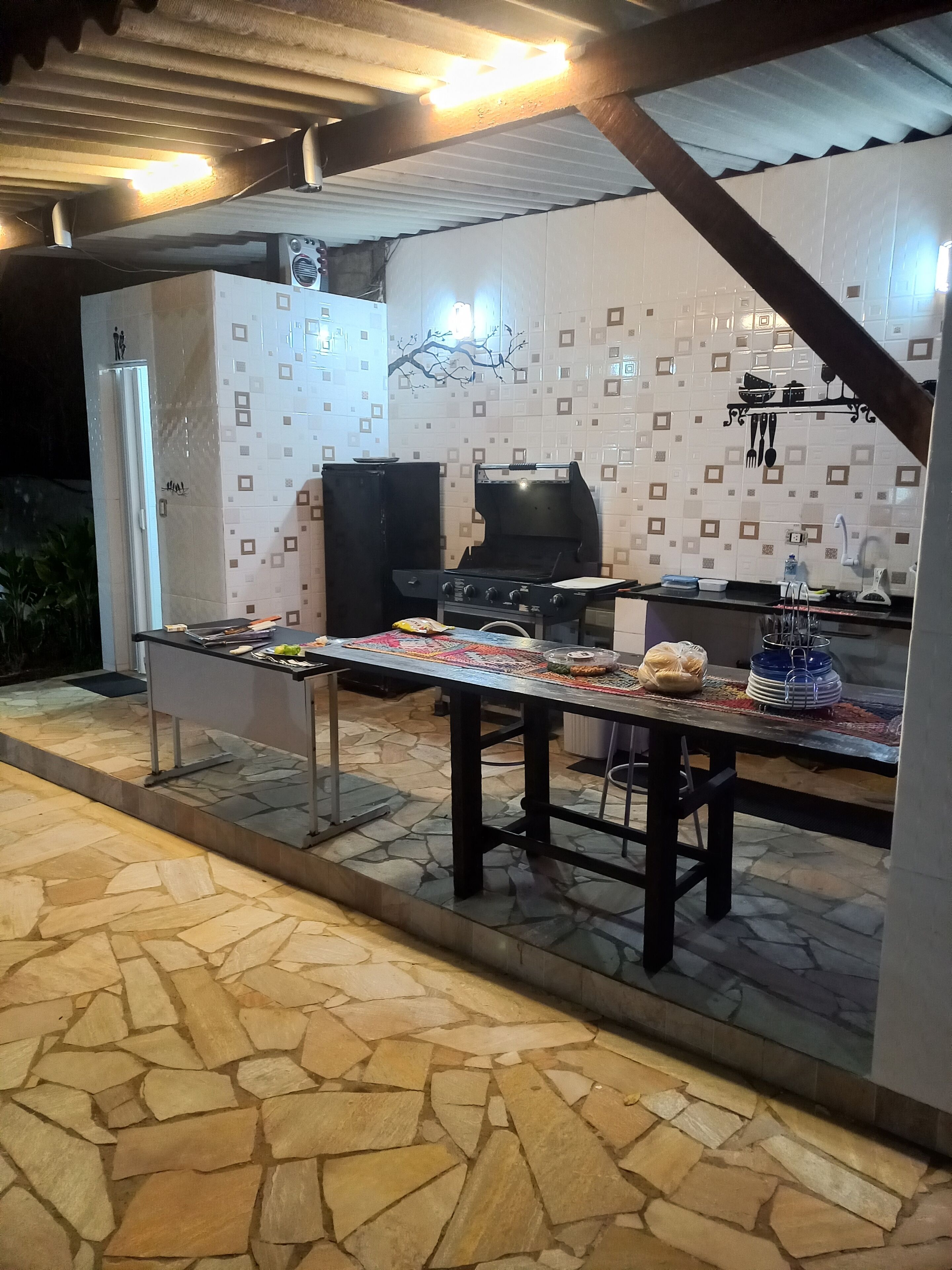 An outdoor kitchen area with a stone-tiled floor, a grill, and a table set with various items.