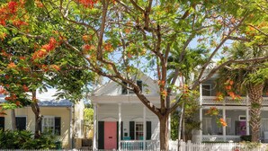 A white house with a porch, surrounded by a white picket fence, and a tree with red flowers. - CONCH HEAVEN ~ Stylish 2B/2Ba, Sleeps 6 in Old Town w/ Heated Cocktail Pool! (Key West)
