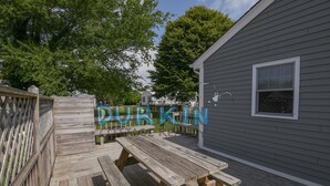 Een houten terras met picknicktafels, een grijs gebouw met een raam en een omheining.