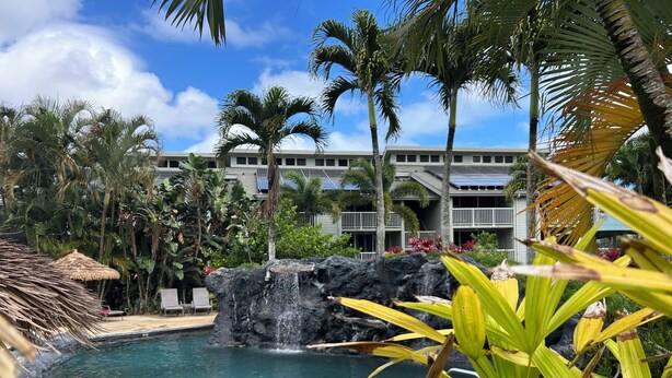A pool with a waterfall, surrounded by palm trees and a building with balconies. - Full Ocean View, Great For Whale Watching (Princeville)