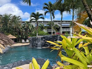 A pool with a waterfall, surrounded by palm trees and a building with balconies. - Full Ocean View, Great For Whale Watching (Princeville)
