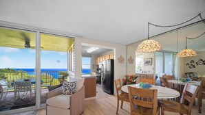 A modern living room with a sofa, dining table, and chairs, a kitchen in the background, and a large sliding glass door leading to a balcony with a view of the ocean. - Full Ocean View, Great For Whale Watching (Princeville)